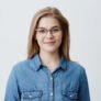 Horizontal portrait of smiling happy young pleasant looking female wears denim shirt and stylish glasses, with straight blonde hair, expresses positiveness, poses against gray background with copy space for promotional text
